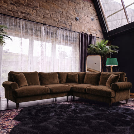 A large L-shaped sectional sofa upholstered in dark brown velvet fabric, styled in a loft-style room with a brick wall, sheer curtains, a dark rug, and a green lamp.