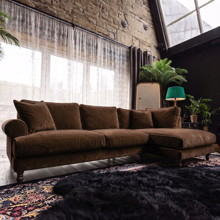 A brown velvet sectional sofa with a chaise lounge, styled in a rustic loft room with a brick wall, sheer curtains, plants, and a dark rug.