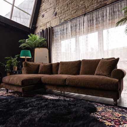 A brown velvet sectional sofa with matching throw pillows, styled in a loft-style living room with a brick wall, sheer curtains, a green lamp, and a dark shag rug.